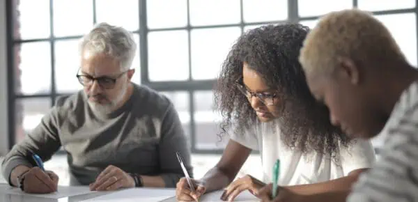 Serious multiethnic colleagues in casual wear writing on papers with pen during teamwork while sitting at table in light office behind fenced window