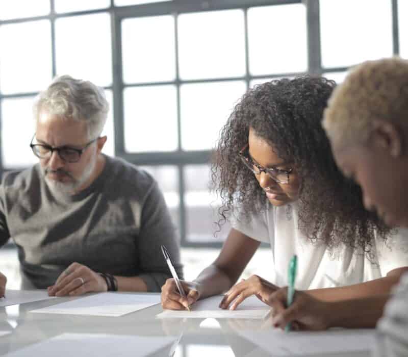 Serious multiethnic colleagues in casual wear writing on papers with pen during teamwork while sitting at table in light office behind fenced window
