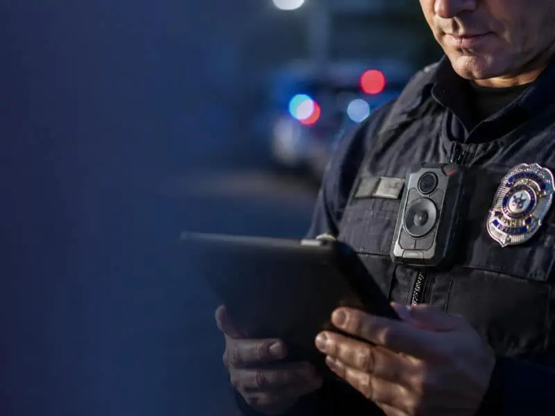 Police officer wearing a body camera and reviewing an AI-generated report on a tablet.
