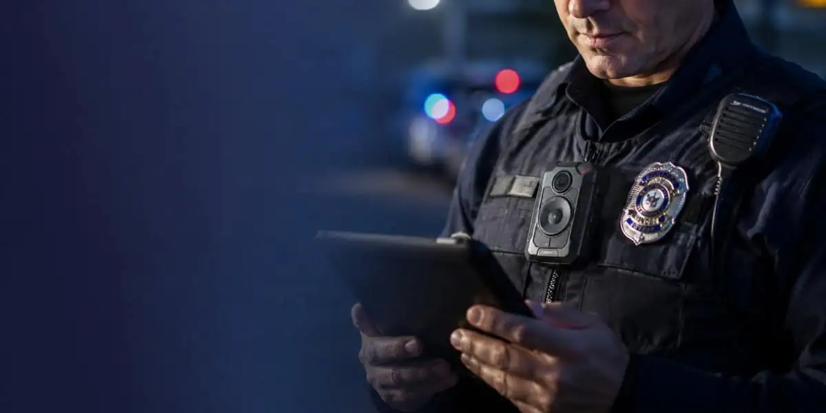 Police officer wearing a body camera and reviewing an AI-generated report on a tablet.
