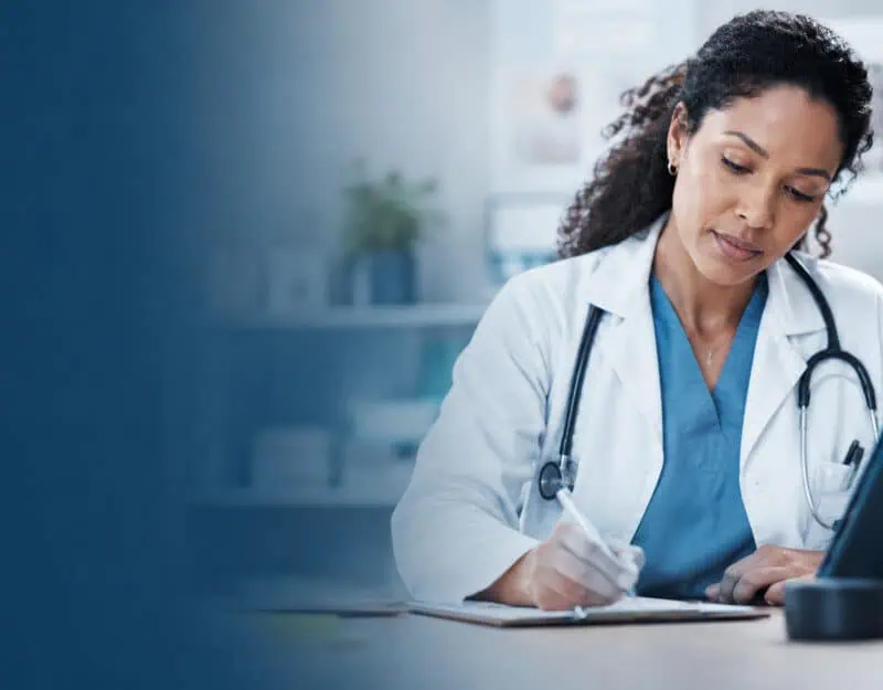 A physician reviews patient information while writing clinical notes beside a tablet in a modern medical office, representing AI-assisted medical documentation and clinical recordkeeping.