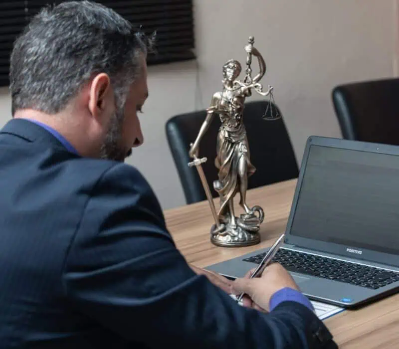 Over-the-shoulder view of an attorney in a dark suit working on a laptop at a desk, with a bronze Lady Justice statue beside him, illustrating AI use in legal work and discovery risk.