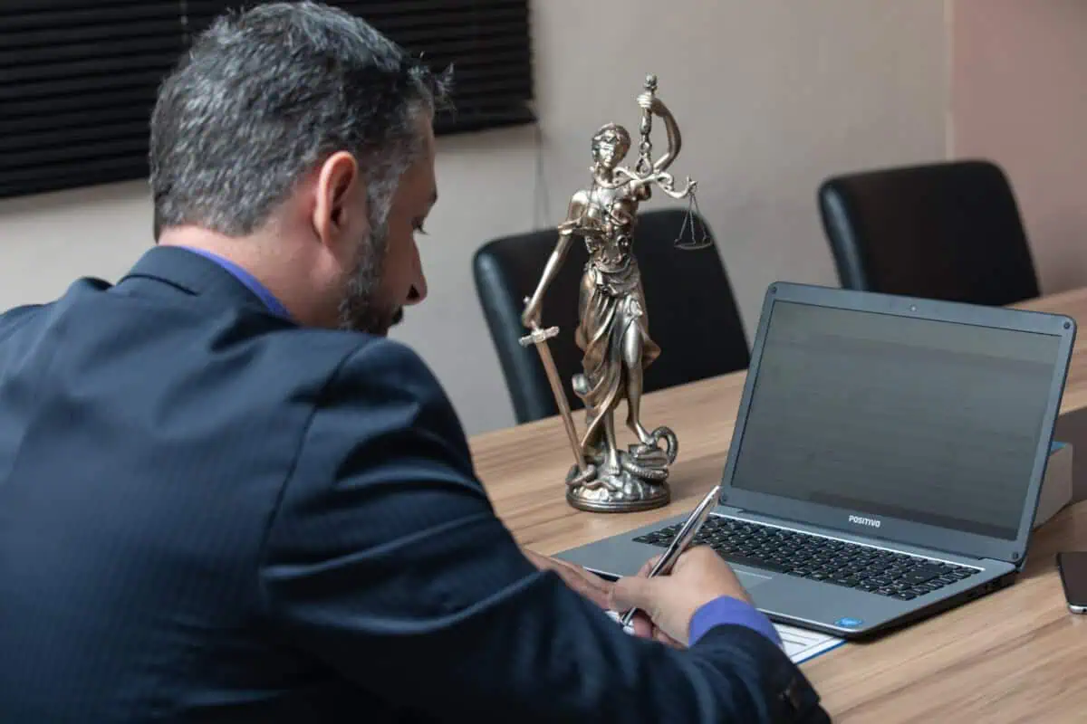 Over-the-shoulder view of an attorney in a dark suit working on a laptop at a desk, with a bronze Lady Justice statue beside him, illustrating AI use in legal work and discovery risk.
